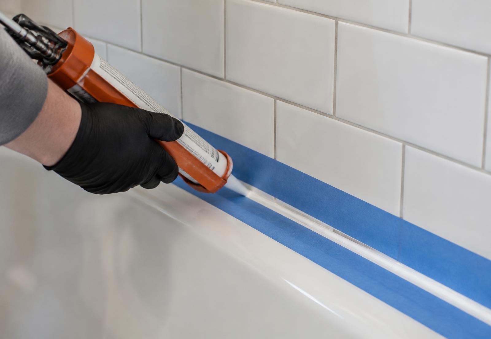 Gloved hand running a caulk bead along a bathtub under subway tile
